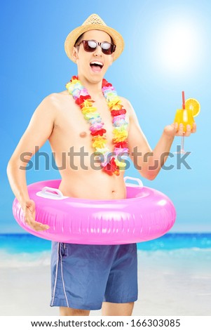 Smiling man in swimming shorts, holding an orange cocktail and posing on a beach next to a sea