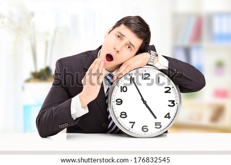 Sleepy young businessman leaning his head on a big wall clock, indoors