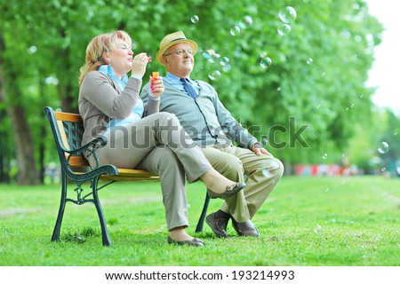 Elderly couple blowing bubbles in park seated on bench