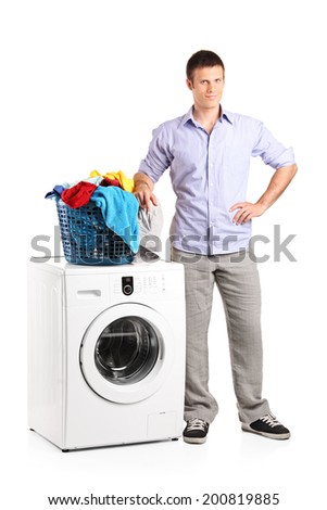 Full length portrait of a guy standing by a washing machine with a laundry basket on it isolated on white background