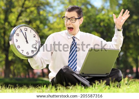 Young angry businessperson with computer sitting on grass and looking at clock in a parK