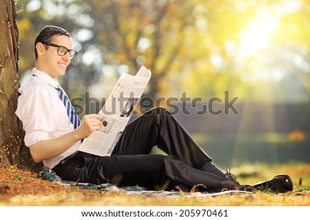 Young man with tie sitting on a grass and reading a newspaper in a park