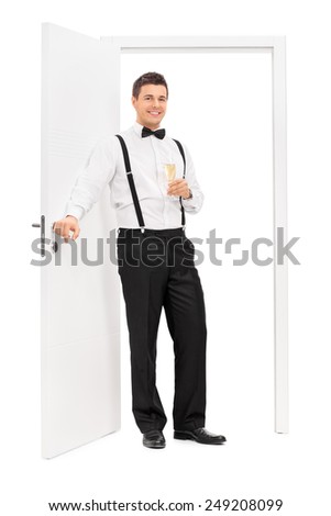 Full length portrait of an elegant guy standing by a door and holding a glass of wine isolated on white background