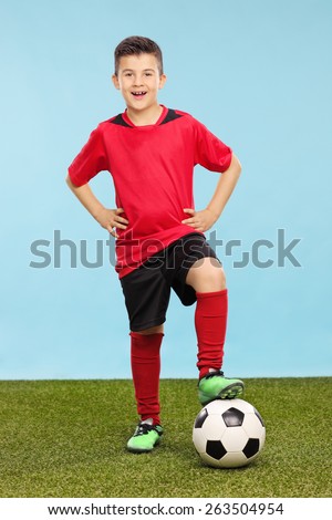 Full length portrait of a junior in a soccer uniform standing over a soccer ball on a grass field with a blue background