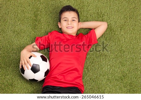 Relaxed youngster in a red football jersey lying on a pitch, holding a football in his hand and looking at the camera