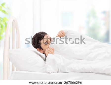 Scared guy lying in bed and hiding under a blanket at home