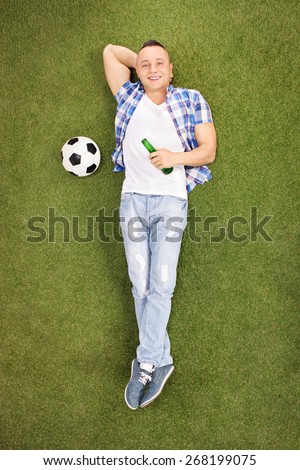 Young guy in casual clothes lying on green grass and holding a bottle of beer, with a soccer ball beside him