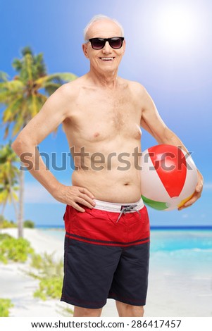 Vertical shot of a senior in swim shorts holding a beach ball at a beach and looking at the camera