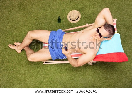 Relaxed young man sitting on a sun lounger and listening to music on his cell phone in his backyard