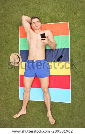 High angle vertical shot of a handsome young man lying on a towel in his backyard and listening to music on his phone