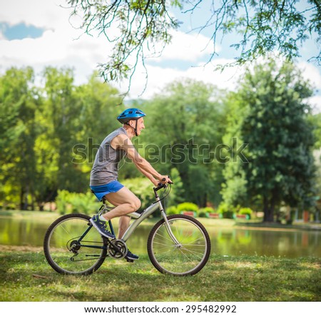Profile shot of a joyful senior biker riding a bicycle in a park by a pond on a beautiful summer day shot with tilt and shift lens