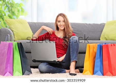Young woman holding a laptop and sitting in front of a couch with a bunch of shopping bags around her at home