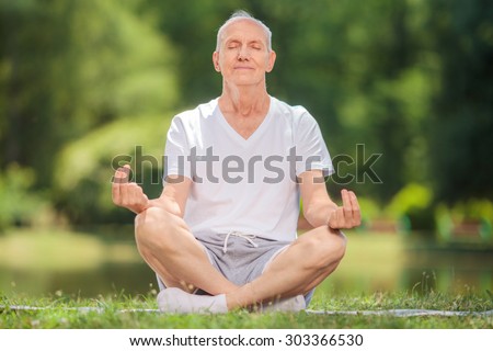 Peaceful senior man meditating seated on a blanket in a park by a lake