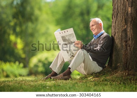 Cheerful senior gentleman reading a newspaper in park and leaning against a tree seated on the ground on beautiful summer day