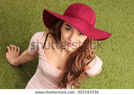 High angle shot of a cheerful young woman with a stylish hat sitting on grass and looking at the camera