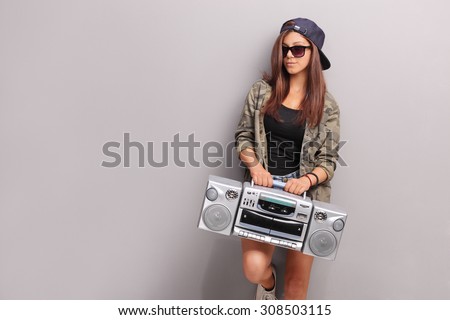 Cool teenage girl in hip hop outfit holding a ghetto blaster and leaning against a gray wall