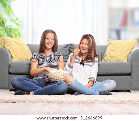 Teenage sisters eating popcorn and watching TV seated on the floor in front of a gray sofa at home
