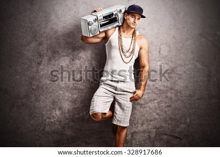 Young male rapper in hip-hop outfit listening to music from a ghetto blaster and leaning against a rusty gray concrete wall