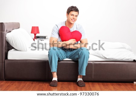 Young man sitting on a bed and holding a heart shaped pillow