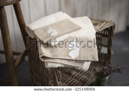 A pair of towels with embroidered flower design on edges atop a rectangular woven box beside wooden stool over wooden panel bakground.