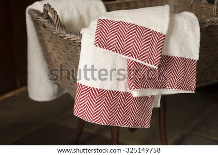 Three white hand towels with maroon herringbone pattern on edges hanging from a woven rectangular basket on a wooden stool.