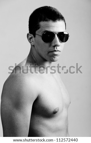 Stunning young man, with a very serious expression, appealing as the bad guy. The man is wearing sun glasses and his muscles are visible. A dramatic lighting has been used.