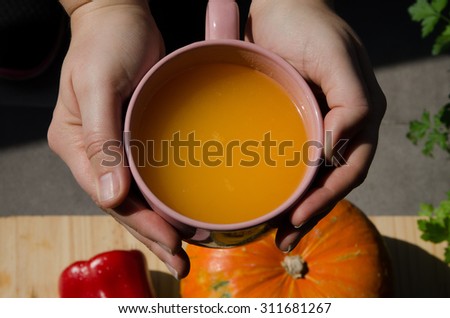 Hands holding a pumpkin soup cup with organic squash and vegetables in the background