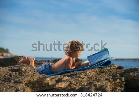 Cute little boy lying on his stomach reading book on the beach. Some negative space around.
