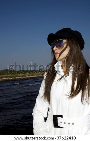 Beautiful brunette fashion model with a white coat, black beret and sunglasses standing by the sea