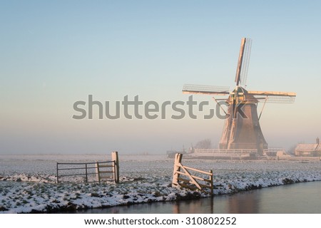 Winter landscape in Holland (the Netherlands) in a rural area with a windmill in a meadow, a fence, a canal, snow and fog at sunrise.