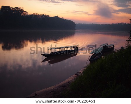 A boat navigating the Tambopata jungle river during a purple and pink sunrise with reflection in the water in the Amazon rainforest in eastern Peru.