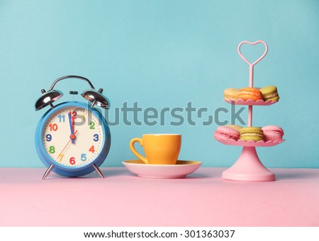 Cup of coffee and alarm clock on the blue and pink background with macarons