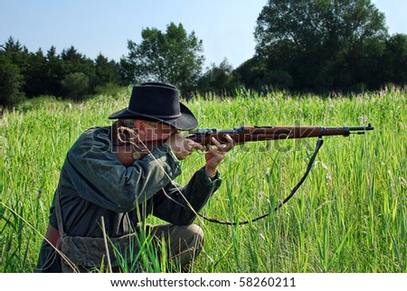 Hunter crouches in the grass and aims his rifle