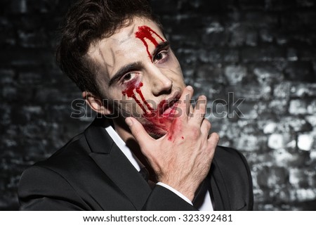 Waist up portrait of a man wearing a suit, looking at the camera frighteningly with zombie bloody make up on his face, wiping blood from his lip, black brick wall on the background