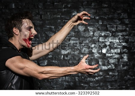 Waist up photo of a young man with bloody make up, standing in a profile shouting and stretching his hands to copy place on the black brick wall