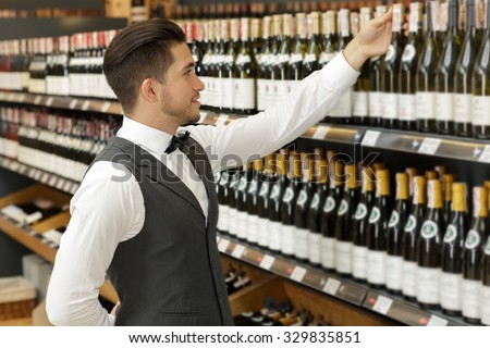 Portrait of a handsome bearded sommelier standing in a shop taking a bottle from the shelf, wearing a suit