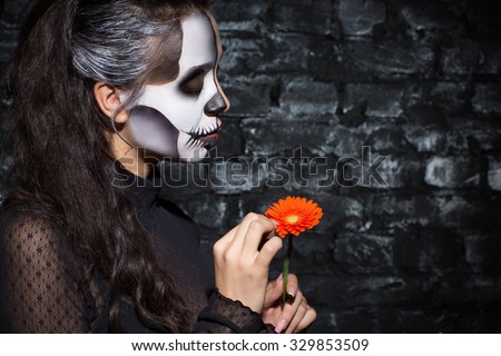 Halloween masquerade party. Portrait of a beautiful woman standing in a profile with a skeleton make up gently holding an orange flower and looking at it