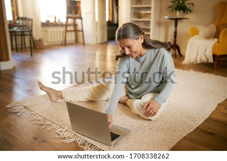 Indoor image of beautiful energetic female on retirement sitting barefoot on floor using laptop turning on calm music for meditation. Elderly European woman surfing internet on portable computer