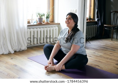 Cheerful attractive young overweight woman in activewear choosing healthy lifestyle, sitting on mat with hands on bare feet, doing butterfly yoga exercise, stretching thighs. Body shape and activity Foto stock © 