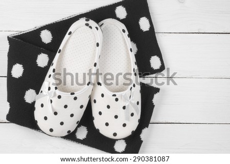 Pair of feminine slippers with white and black polka dot decor and woolen scarf on white wooden floor.