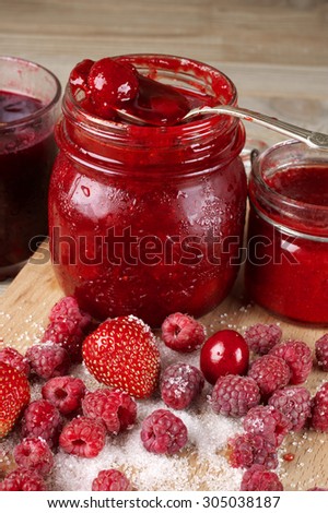 Assorted jams in glass jars and fresh berries in sugar on wooden cutting board.