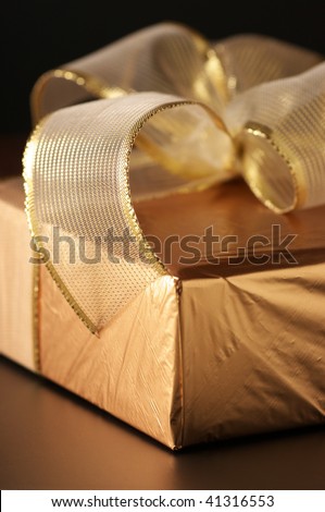 Close-up of gold foil gift with gold translucent bow on black background. Selective focus on front edge of ribbon.