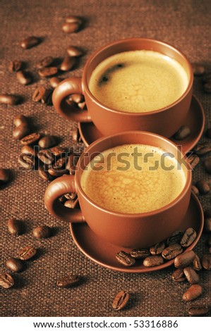 Two brown ceramic cups of coffee with froth and roasted coffee beans on brown canvas.