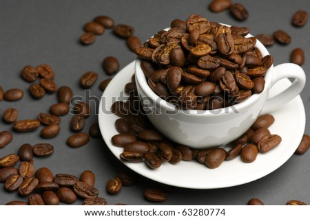 Roasted coffee beans in white cup with saucer on dark gray background.