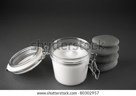 Glass jar of cosmetic cream and stack of massage stones on dark grey background.