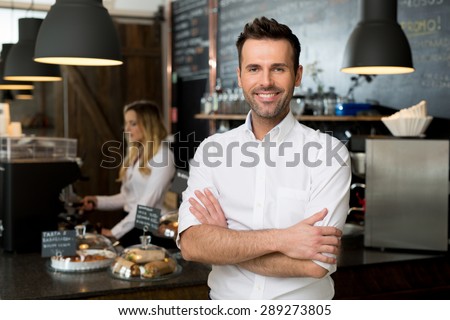 Successful small business owner standing with crossed arms with employee in background preparing coffee