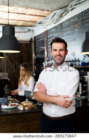 Successful cafe owner standing with crossed arms with employee in background preparing coffee