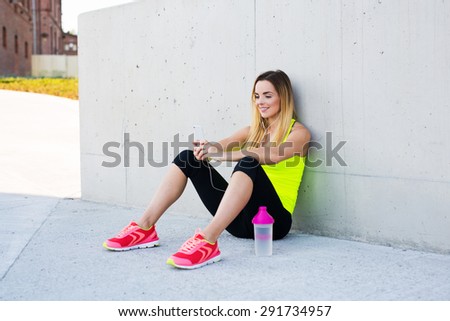 Female runner resting after workout, holding smartphone in her hands and listening to music
