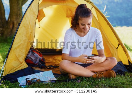 Happy young woman sitting outside tent with smartphone