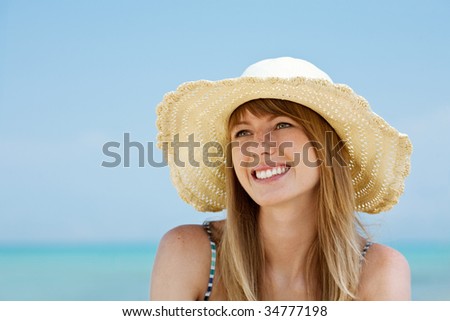 Beautiful young woman with sun hat sitting and smiling on the beach. Sea in background.